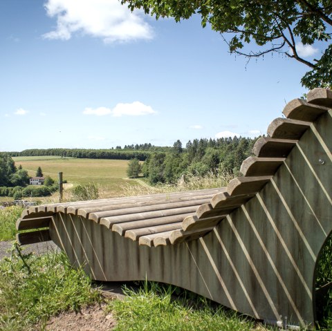 Holzliegebank mit Blick auf grüne Felder und Wälder unter blauem Himmel in Neidenbach., © TI Bitburger Land - Monika Mayer