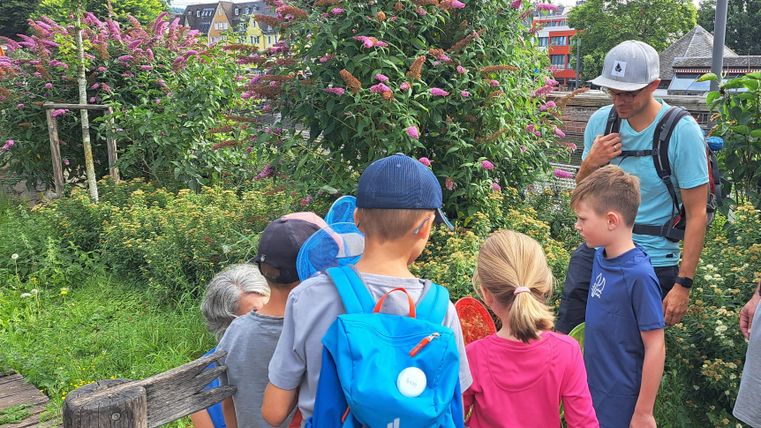 Eine Gruppe von Kindern steht in einem Garten und beobachtet Pflanzen. Ein Erwachsener erklärt etwas zu den Pflanzen im Hintergrund.