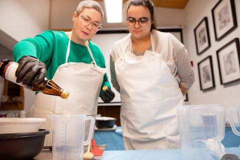 Two women in aprons are working in a kitchen. One woman is pouring a liquid into a measuring cup.