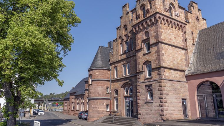 Een historisch gebouw in de neogotische stijl met torens en rode bakstenen. Op de achtergrond zijn bomen en een helder blauwe lucht te zien.