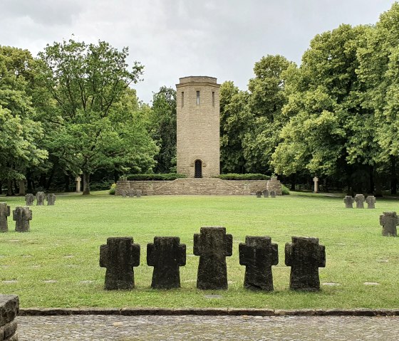 Erebegraafplaats Kolmeshöhe in Bitburg: een centrale toren omringd door een groene weide en verschillende stenen kruisen, ingebed in een bos.