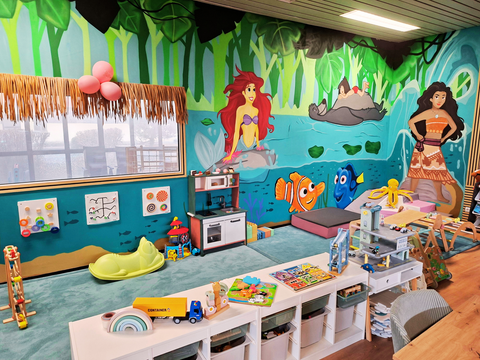 Een kleurrijke speelruimte met vrolijke muurschilderingen, waaronder figuren uit Disney-films. Er zijn speelgoed, een keuken en gezellige zitplaatsen voor kinderen.