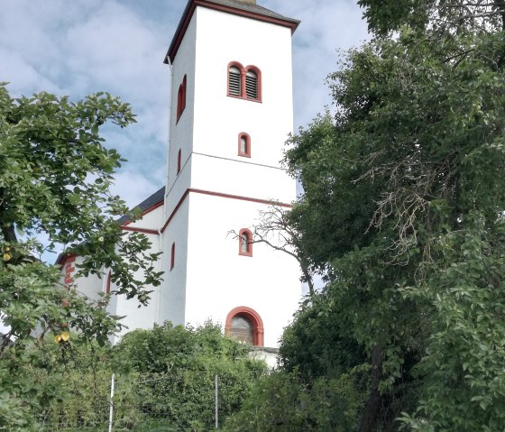 Une église blanche avec un toit et une tour rouges, entourée d'arbres verts et d'un ciel bleu en arrière-plan., © TI Bitburger Land