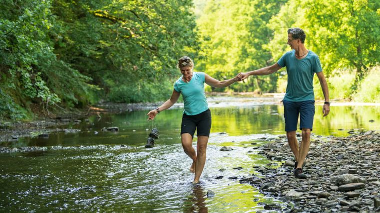 Een stel loopt op blote voeten door een ondiepe rivier, omgeven door groene natuur.