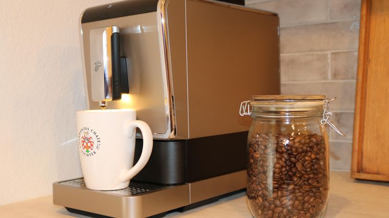 Une cafetière moderne est posée sur un plan de travail à côté d'un verre contenant des grains de café. Une tasse blanche se trouve sous le bec de la machine.