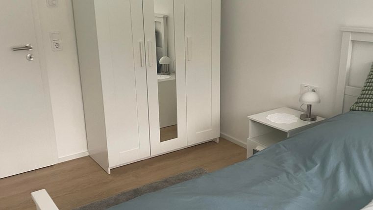 Ein helles Schlafzimmer mit einem weißen Kleiderschrank und einem Bett mit blauen Bettwäsche. Neben dem Bett steht ein Nachttisch mit einer Lampe.