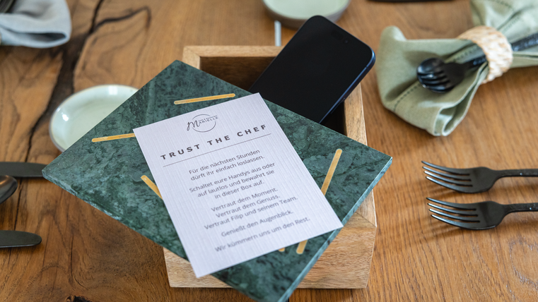 Une table élégante avec un plateau en bois, sur laquelle se trouvent un menu vert et un smartphone. À côté du menu, des couverts et des serviettes sont disposés de manière décorative.