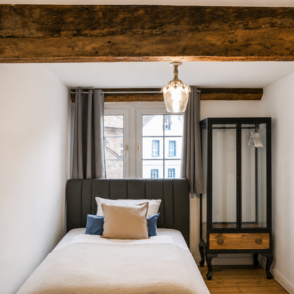 A cozy room with a single bed and a wooden shelf. The window offers a view of a quiet street and the room has a warm, inviting atmosphere.