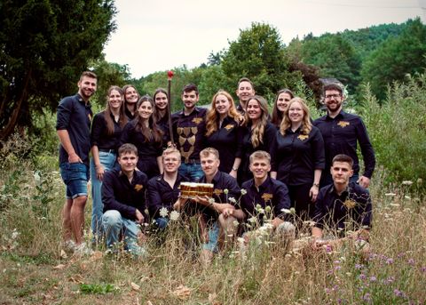 A group of young people is smiling on a meadow. They are giving thumbs up and presenting a tray of drinks.