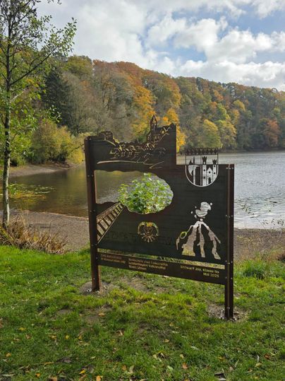 Ein hölzernes Schild am Ufer eines Sees, umgeben von buntem Herbstlaub. Im Hintergrund sind Bäume und ein ruhiges Gewässer zu sehen.