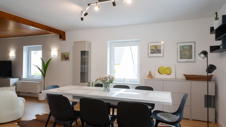 Ein modernes Esszimmer mit einem großen weißen Tisch und schwarzen Stühlen. Die Wände sind hell gestrichen und mit Kunstwerken dekoriert.