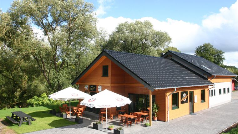 Houten gebouw met terras, parasols en zitgelegenheden, omringd door bomen en een blauwe lucht.