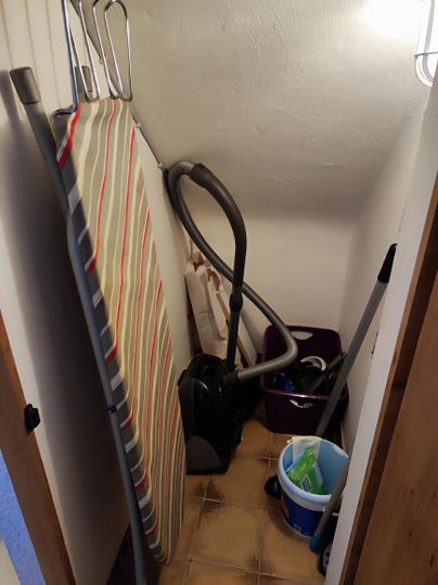In the storage room, there is an ironing board, iron, vacuum cleaner, and cleaning supplies.