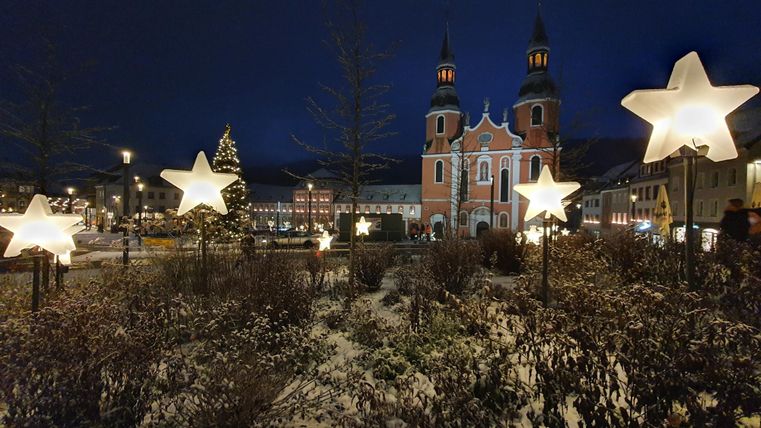 Een indrukwekkende kerk met twee hoge torens ligt voor een schilderachtige achtergrond. Omringd door historische gebouwen en bomen strekt zich een royaal plein uit.