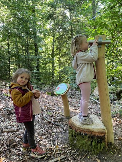 Zwei Kinder im Wald, eines schaut durch ein Holzfernrohr, das andere steht daneben.