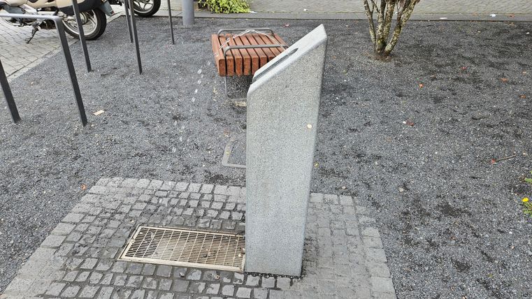 Une pierre grise avec une surface inclinée se trouve dans une zone pavée. À l'arrière-plan, quelques bancs et arbres sont visibles.