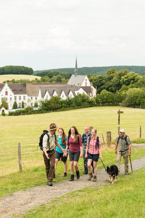 A group of hikers is walking along a path, accompanied by a dog. In the background, there is a beautiful, older building and green fields visible.
