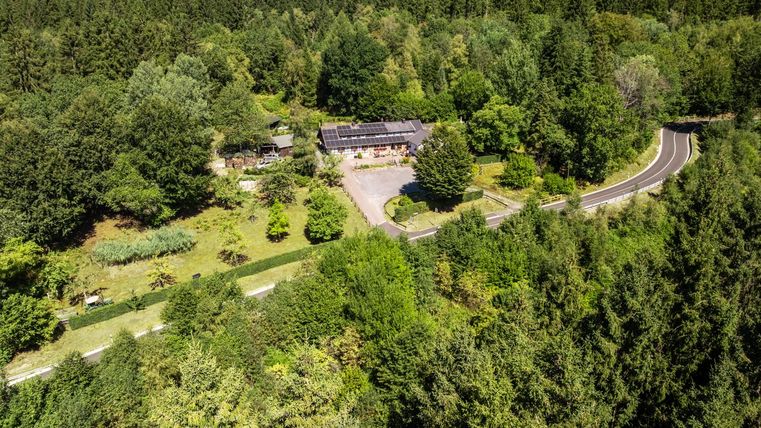 A quiet landscape with a large, surrounding forest and a road that winds through nature. In the foreground, a building with a greened property can be seen.