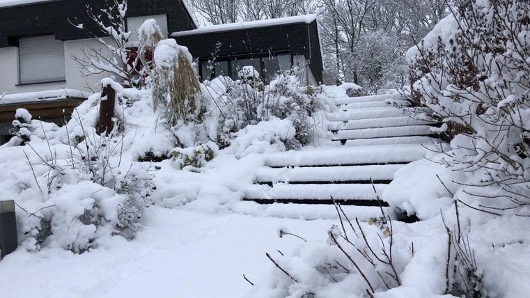 Ein verschneiter Garten mit Treppen, die zu einem Haus führen. Die Landschaft ist winterlich und ruhig.