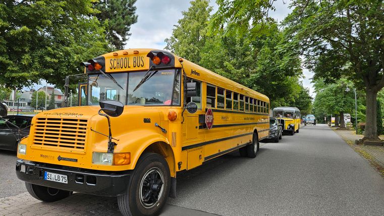 A yellow school bus is parked on a street, surrounded by trees. In the background, more vehicles can be seen.