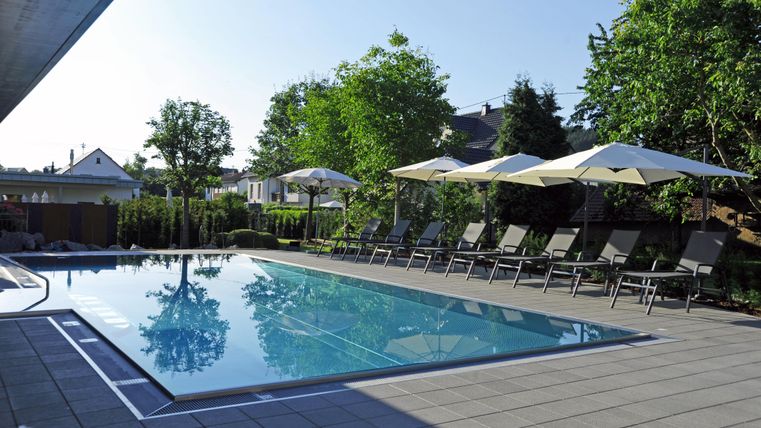 Een heldere pool omgeven door ligstoelen en parasols. De omgeving is groen met bomen en een heldere lucht.