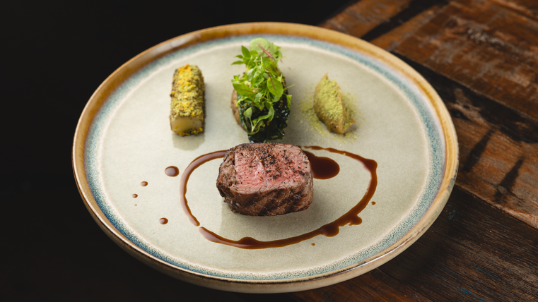 Un plat artistique sur une assiette, qui montre un morceau de viande avec une sauce sombre. Il y a aussi des légumes et des herbes fraîches.