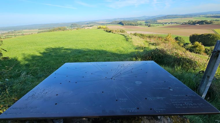 A viewing platform with a large metal panel is visible. In the background, a vast landscape stretches out with green meadows and gentle hills.