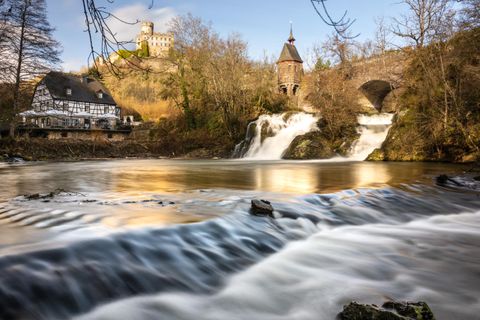A tranquil river with gentle waterfalls flows in front of an old castle. Surrounded by trees and a historic building, the scene is idyllic and picturesque.