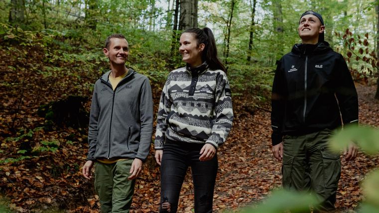 Drie mensen wandelen door een groene bos met herfstbladeren. Ze lachen en genieten van de natuur.