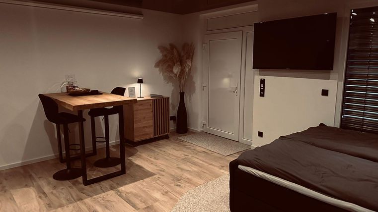 Une chambre moderne avec un lit et un petit coin repas. De grandes fenêtres et des couleurs chaudes créent une atmosphère chaleureuse.