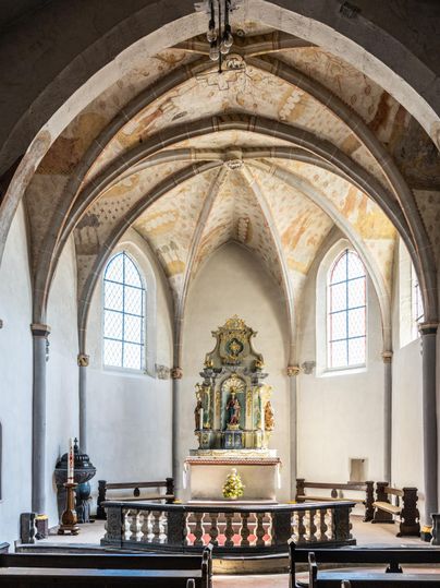 Het interieur van een kerk met hoge, gewelfde plafonds en kleurrijke ramen. In het altaargebied bevindt zich een kunstzinnige standbeeld.