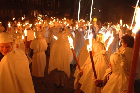 Eine Gruppe von Menschen in weißen Gewändern feiert in der Nacht mit brennenden Fackeln. Die Szene strahlt eine mystische und festliche Atmosphäre aus.