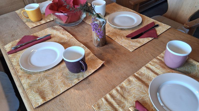 Een mooi gedekte tafel met borden, kopjes en kleurrijke servetten. De tafelkleed is geel met een bladerenpatroon.