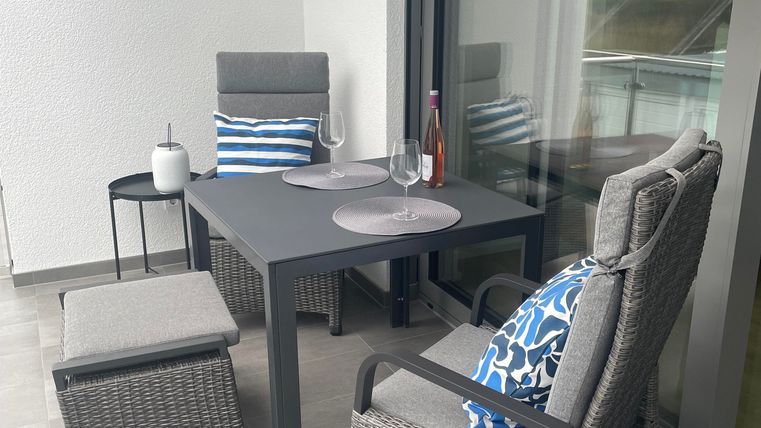 Ein gemütlicher Balkon mit einem Tisch und Stühlen aus Rattan. Es gibt ein paar Accessoires und einen Blick auf die Umgebung.