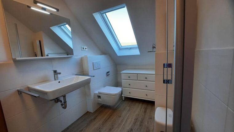 Ein modernes Badezimmer mit Fenster und hellem Holzfußboden. Es gibt ein Waschbecken, eine Toilette und einen Schrank.