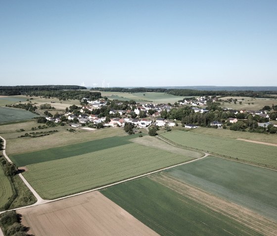 Vue aérienne d'Idenheim, entourée de champs verdoyants. En arrière-plan, on aperçoit plusieurs éoliennes qui s'étendent à l'horizon., © Ingrid Penning