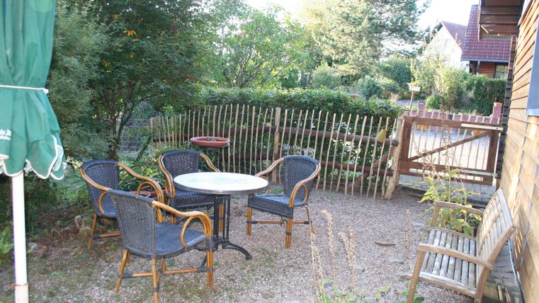 Een gezellige buitenruimte met een ronde tafel en stoelen van riet. Omringd door groene planten en een houten hek.