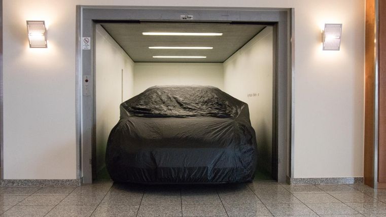 Une voiture recouverte est dans un ascenseur. Les murs sont clairs et le sol est carrelé.