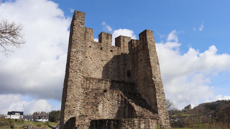 An old stone fortress with four towers and an impressive structure. In the background, green meadows and a cloudy sky can be seen.