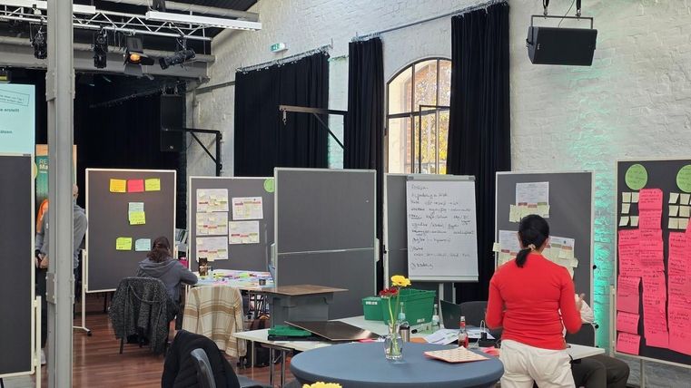 Een creatieve werkruimte met verschillende standen en gekleurde post-its. Mensen zijn bezig om ideeën te ontwikkelen en te bespreken.