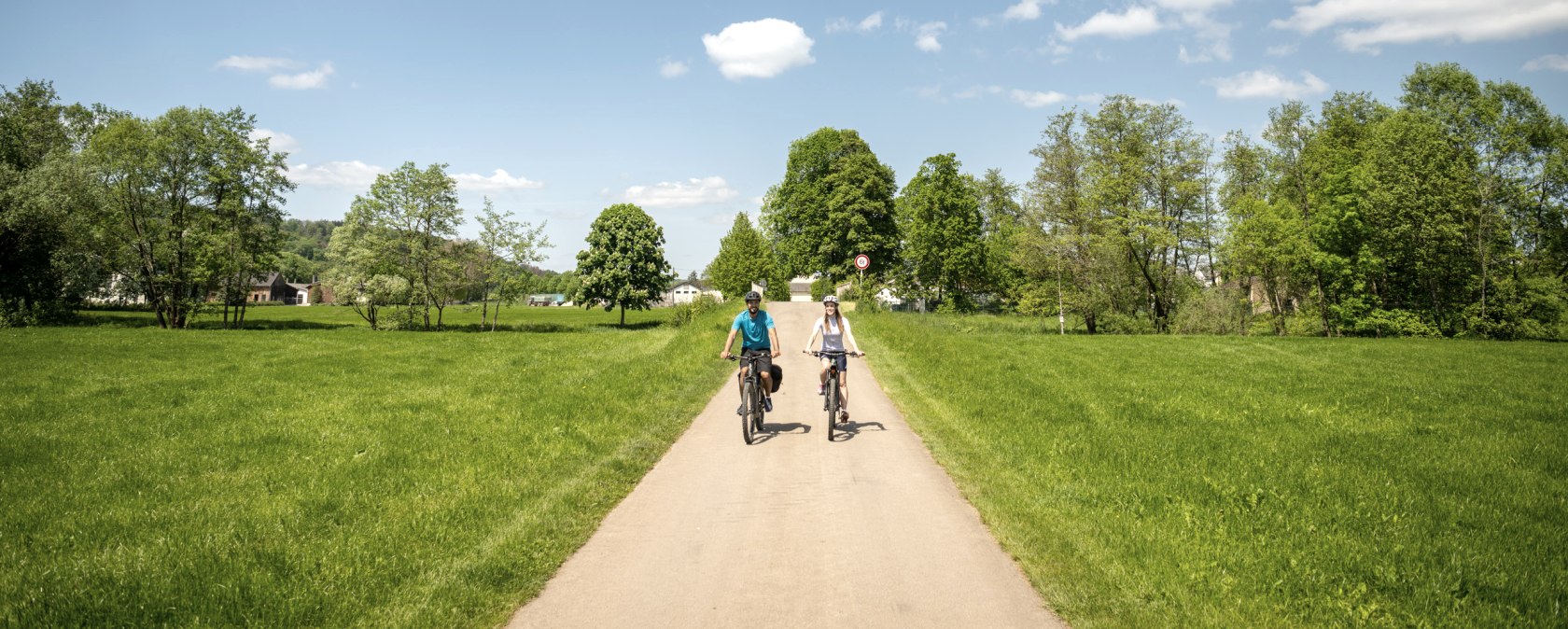 Zwei Radfahrer fahren auf einem asphaltierten Weg durch eine grüne Landschaft. Der Himmel ist blau mit einigen Wolken., © Eifel Tourismus GmbH, Dominik Ketz
