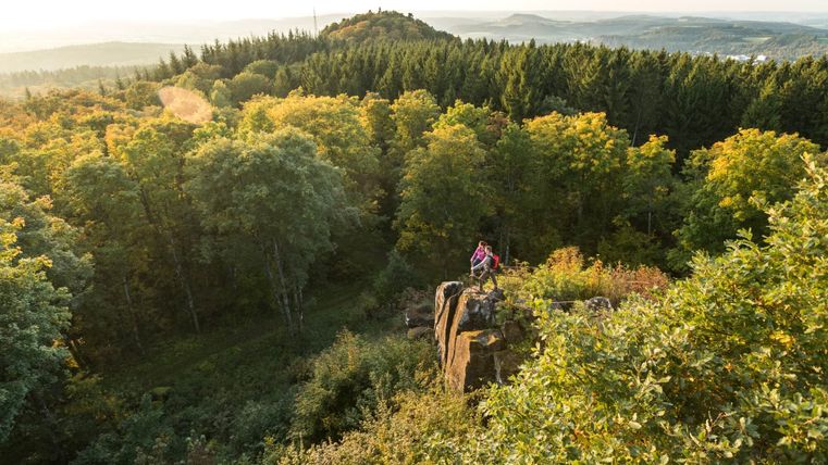 Deux personnes se tiennent sur un rocher avec vue sur une forêt et des collines en arrière-plan.