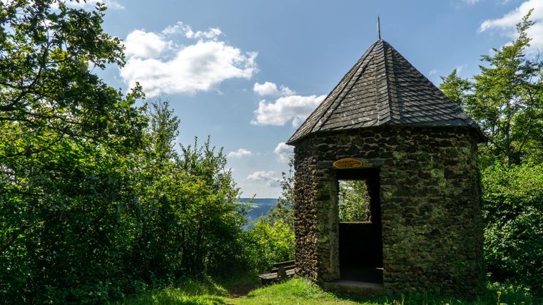 Ein steinernes Pavillon mit spitzem Dach steht in einer grünen Landschaft unter blauem Himmel.