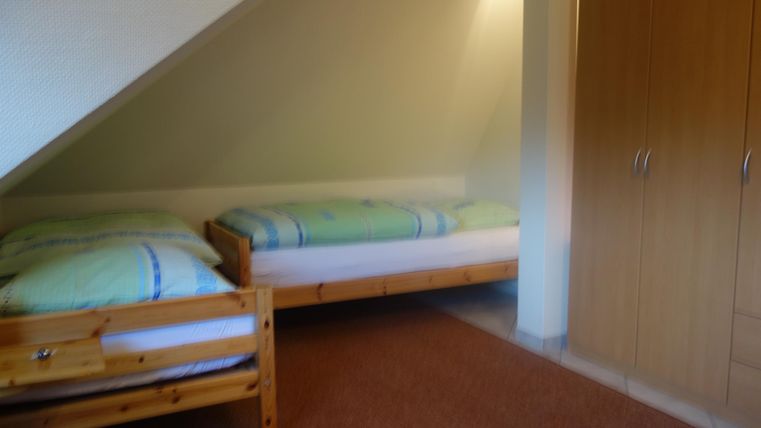 A simple bedroom with two single beds and a wooden wardrobe. The walls are light and the floor is carpeted.