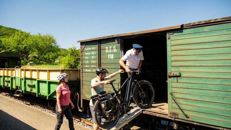 Ein Mann trägt sein Fahrrad in einen begrünten Waggon, während eine Frau zuschaut. Es ist ein sonniger Tag mit blauen Himmel und viel Grün im Hintergrund.