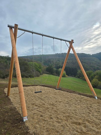 Eine Schaukel aus Holz steht auf einem Spielplatz in der Natur. Im Hintergrund sind sanfte Hügel und bewaldete Flächen zu sehen.