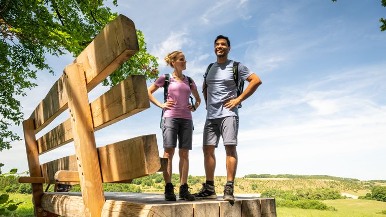 Zwei Personen stehen auf einer großen Holzbank in einer grünen Landschaft.