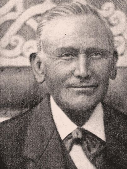 An older man in a suit and tie smiles at the camera. The background shows a simply decorated wall.