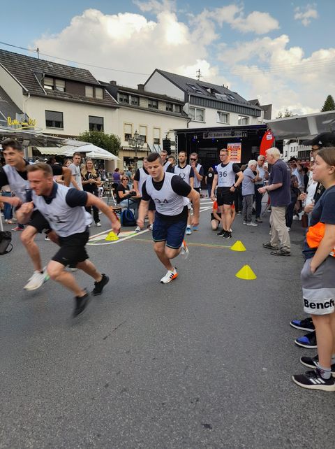 Eine Gruppe von Männern nimmt an einem Wettlauf teil. Im Hintergrund stehen Zuschauer und einige Stände.