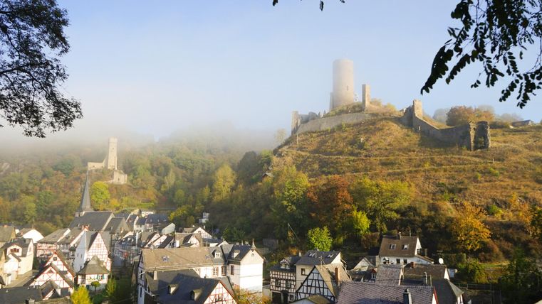 Eine malerische Landschaft mit einer alten Burg auf einem Hügel und mistiger Umgebung. Darunter liegt ein charmantes Dorf mit traditionellen Häusern.